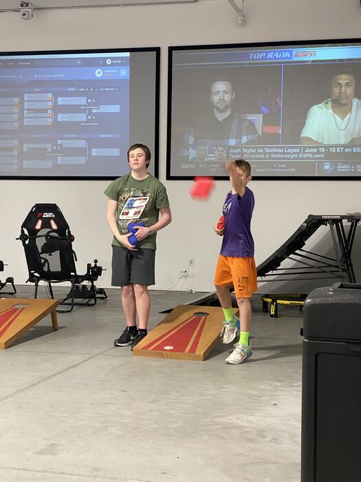Two teens playing cornhole with the live tournament bracket on the big screen